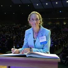 Portrait of Nancy Makri on stage in a large auditorium signing a book during an induction ceremony for the American Academy of Arts and Sciences with audience in the background.