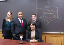 Photo of the four researchers in front of a blackboard with three standing and Professor White sitting next to an hourglass.