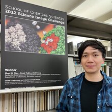 Chun Kit Chan stands next to a poster of his science image that is hanging on a book shelf in the chemistry library