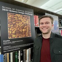 Nathan Forney stands next to a poster of his science image hanging on a bookshelf in the chemistry library.