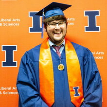 Portrait of Ian Garvey in graduation regalia standing in front of an orange background with blue Block I logos