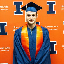 Portrait of Matthew Dake in graduation regalia standing in front of an orange background with blue Block I logos