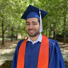 Portrait of Qasim Sikander in graduation regalia standing in front of entrance to Noyes.
