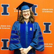 Portrait of Sophie McClain in graduation regalia standing in front of an orange background with blue Block I logos