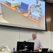 Greg George talks in a lecture classroom displaying some of the equipment he has worked with on a large overhead screen.