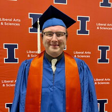 Zach Burke standing in graduation regalia in front of an orange backdrop