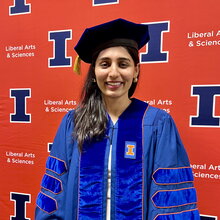 Saman Fatima stands in graduation regalia in front of orange backdrop