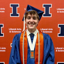 Joel Ryan stands in graduation regalia in front of orange backdrop