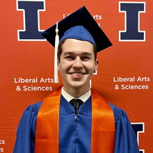 Ethan Ramirez stands in graduation regalia in front of orange backdrop