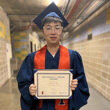 Zikang Xu stands in graduation regalia in front of orange backdrop