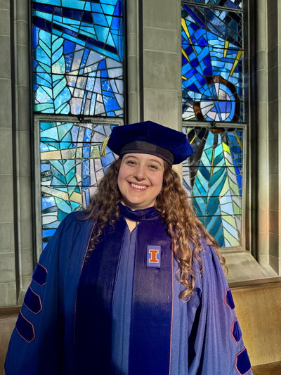 Katie Hatzis standing in cap and gown in front of stained glass windows