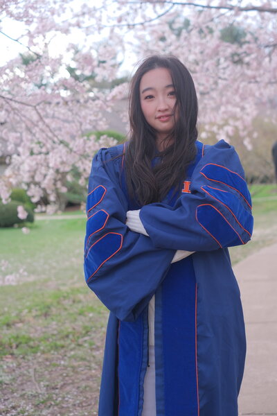 Yanqi Tan standing with arms crossed in graduation robe in front of blooming tree branches.
