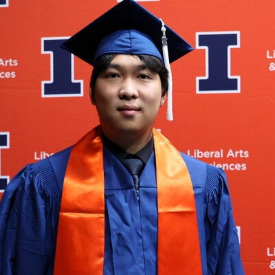 Yi Yang in graduation regalia in front of an orange Block I banner.