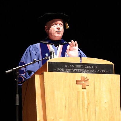 Christopher Cramer in graduation regalia speaking at a podium
