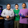 Angad Mehta and two other researchers stand side by side in one of their lab spaces with equipment behind them and holding equipment used in their study. 