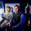 Two researchers in a dark lab sit in front of a refrigerator that stores research items. 