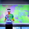 A person wearing a virtual gaming headset and hand controls faces forward in front of a big screen showing the images he is seeing in the headset. 