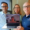 Three people standing side by side holding a laptop with image of a person