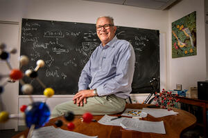 Chemistry professor Thomas Rauchfuss sitting in a classroom in front of a chalkboard with equations written on it.