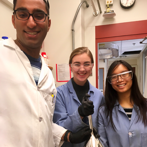 Picture of three chemistry students in lab coats standing together in a lab holding a piece of chemistry equipment. 