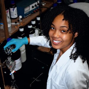 Portrait of  Enleyona Weir in a lab wearing a white lab coat and blue gloves holding a bottle of liquid. 