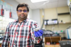 Saket Bhargava stands in the lab holding in one hand a flow electrolysis cell that is a small silver and metal box with two white wires extending from two sides of the box. .