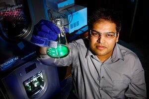Angad Mehta holds a glass container with bright green substance in a dark lab room with equipment behind him. 