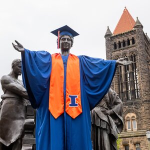 Photo of the Alma Mater dressed in UIUC graduation regalia with Altgeld Hall tower in the background.