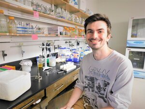Graduate student Jason Cournoyer sits on a chair in a lab next to a bench and shelves filled with chemistry supplies/equipment.