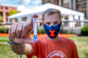 A student standing outdoors on the UIUC campus with a COVID testing tent site in the background holds a tube with blue liquid at the bottom, like the tubes used for collecting COVID saliva samples.