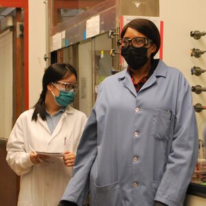 Jeanne N'Diaye in blue lab coat and safety mask and goggles stands the lab teaching electrochemistry to grad students and postdocs.