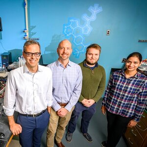 Photo of researchers standing side by side in the lab with blue wall behind them and an all-white-colored molecular structure projected onto the wall.