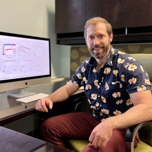 Professor Ben Snyder sits in a desk chair at his office desk with a monitor to his right side showing graphs from his latest research.