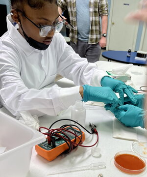 A middle school student wearing a white lab coat and turquoise lab gloves sits at a table working on building a solar cell.