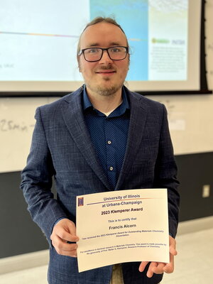 Francis Alcorn stands holding the Klemperer Award certificate in a classroom with a large overhead screen in the background