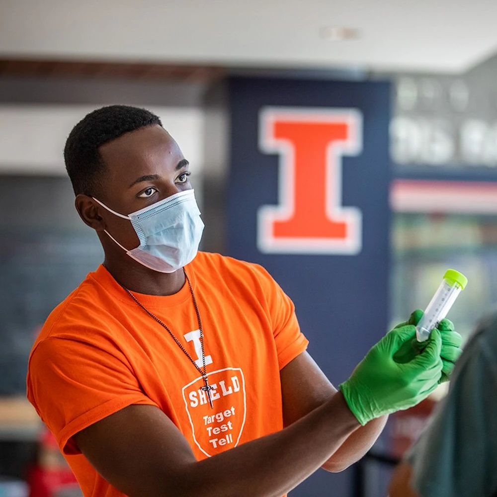 A student volunteer wearing an orange SHIELD team shirt and a mask and rubber gloves holds a saliva testing tube at a COVID-19 testing site on campus. In the background over the student's shoulder is the block I on a placard.