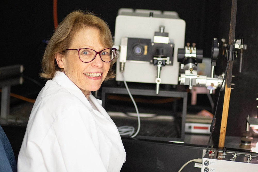 Photo of Deborah Leckband in a white lab coat, turning her face toward the camera while seated in front of lab equipment with a dark background.