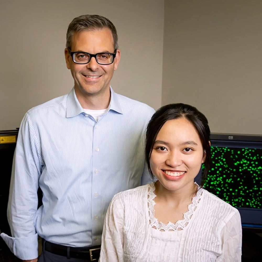 Portrait picture in the lab against tan wall showing Martin Burke standing to the left and behind graduate student Stella Ekaputri, who is sitting.