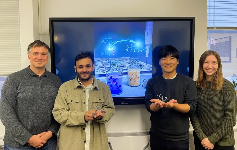 Four people standing in front of a video screen showing a scientific illustration