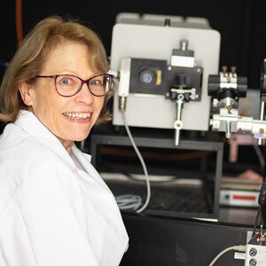 Photo of Deborah Leckband in a white lab coat, turning her face toward the camera while seated in front of lab equipment with a dark background.