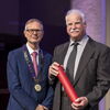 Two men standing side by side in suits and ties with one holding a red certificate tube