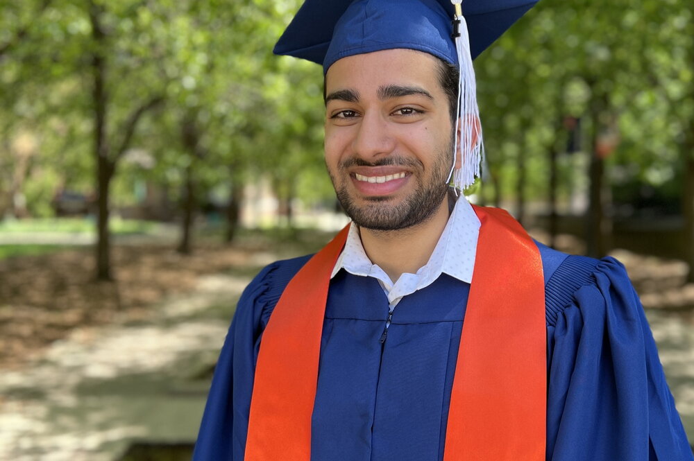 Qasim Sikander in cap and gown outdoors on the UIUC campus with trees in background