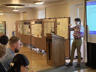 Aidan Lindsay stands at a podium at the front of a room explaining research to an audience.
