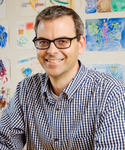 Portrait of Martin Burke seated with a white wall behind him that is full of colorful drawings