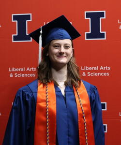 Lucia Urbain in graduation regalia in front of an orange Block I banner.