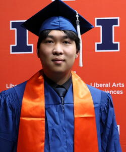 Yi Yang in graduation regalia in front of an orange Block I banner.
