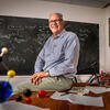 Chemistry professor Thomas Rauchfuss sitting in a classroom in front of a chalkboard with equations written on it.