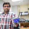 Saket Bhargava stands in the lab holding in one hand a flow electrolysis cell that is a small silver and metal box with two white wires extending from two sides of the box. .