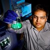 Angad Mehta holds a glass container with bright green substance in a dark lab room with equipment behind him. 