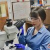 Graduate student Kerry Xu works in a lab wearing a blue lab coat, safety glasses and purple gloves. 